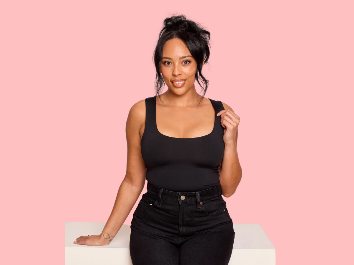 Woman standing confidently against a soft pink background wearing a fitted black sleeveless top and black jeans. She is facing the camera with a relaxed smile, one hand resting on a white plinth and the other lightly holding her top strap. Her dark hair is styled in a loose updo with soft strands framing her face. The image conveys confidence, balance, and natural-looking results, reflecting the goals many patients seek through breast lift surgery, mastopexy, or a natural breast lift.