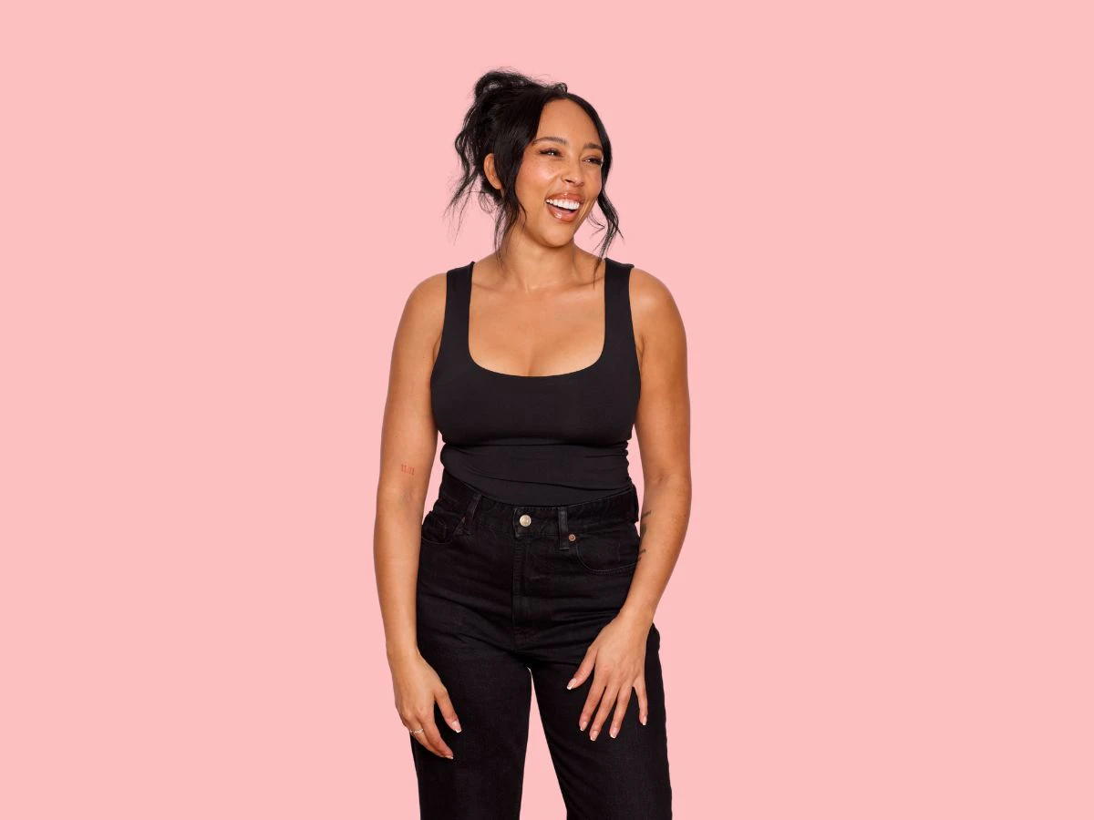 Woman standing against a soft pink background wearing a fitted black sleeveless top and high-waisted black jeans. She is smiling warmly and looking off to the side with a relaxed, confident posture. Her dark hair is loosely styled up with soft strands framing her face. The image conveys confidence, comfort, and body positivity, reflecting the type of result many patients hope to achieve following breast reduction surgery or a breast reduction and lift.